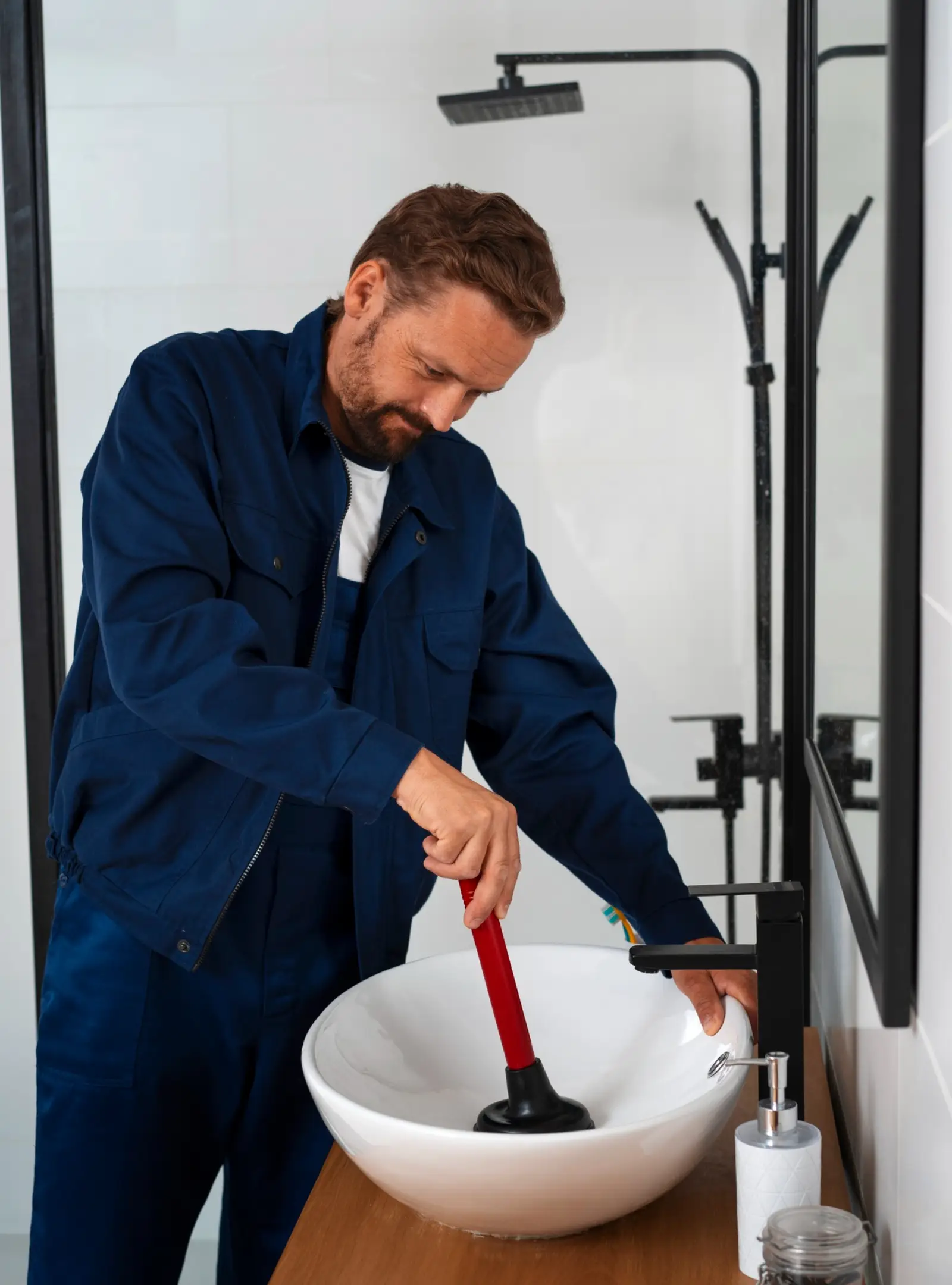 A person in a dark jacket uses a red plunger on a white sink in a modern bathroom setting